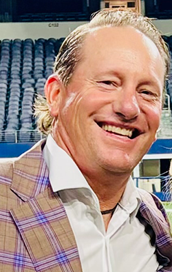 Smiling man in a patterned blazer and white shirt, against a backdrop of stadium seating.