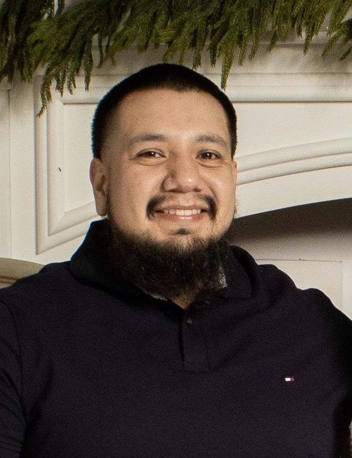 A smiling man with a trimmed beard, wearing a navy blue shirt, against a backdrop of white trim and greenery.