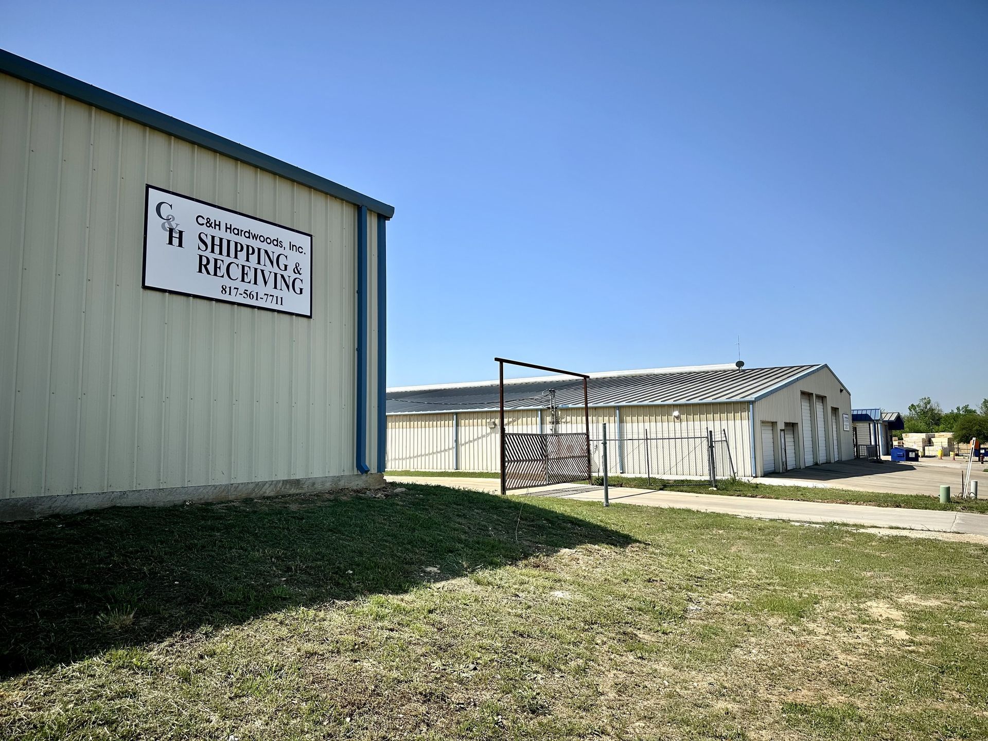 Two warehouses with signs, under a bright blue sky. Green grass and a black gate are in the foreground.