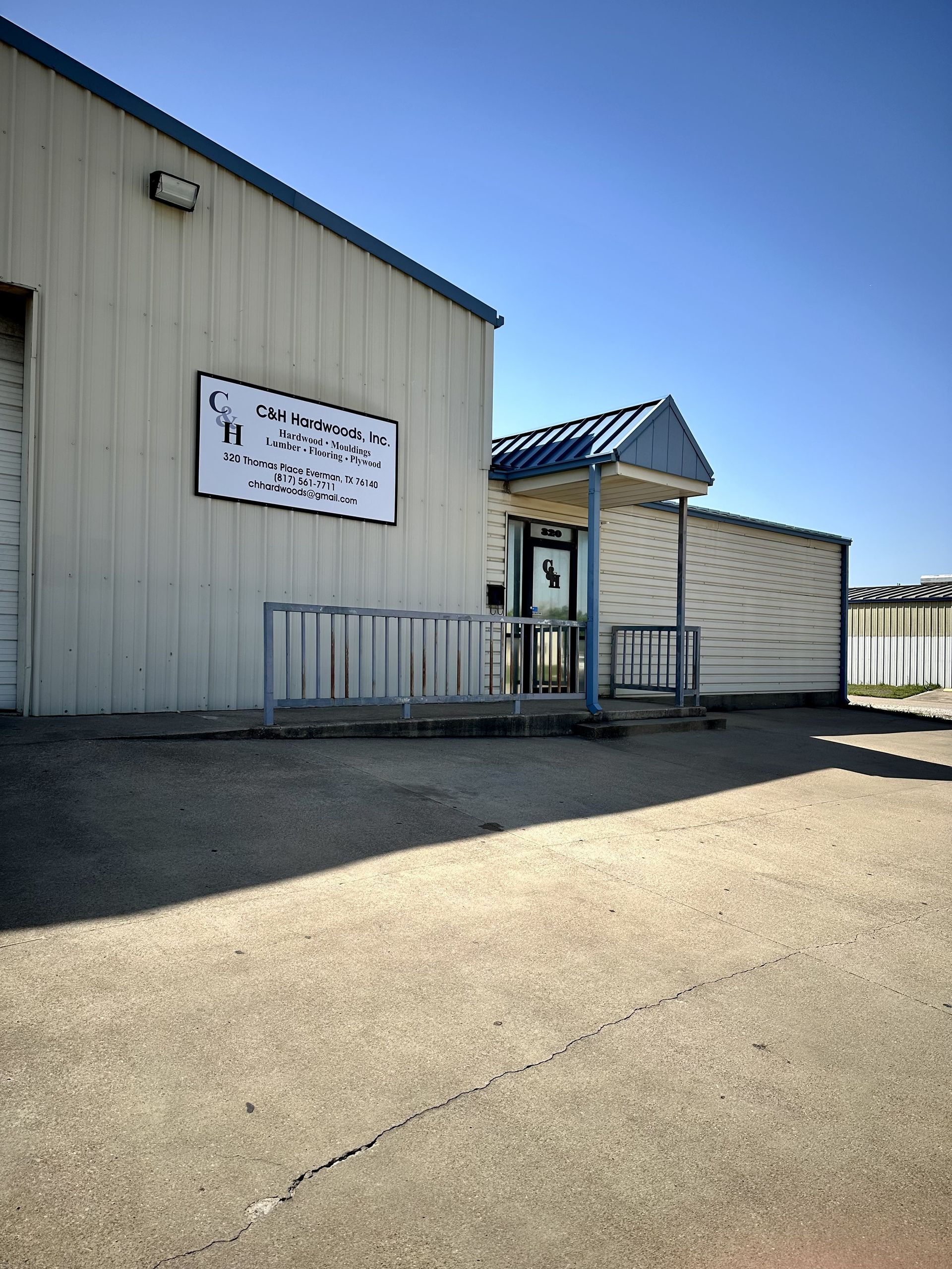 Exterior of a light-colored building with a sign and a small entrance with a blue canopy. The building sits on a paved area under a clear sky.