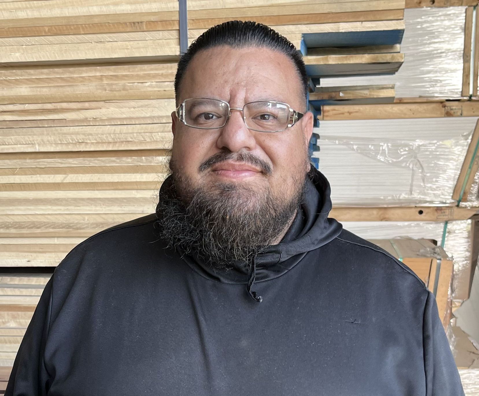 Man with a beard and glasses wearing a black hoodie, standing in front of stacks of wood.