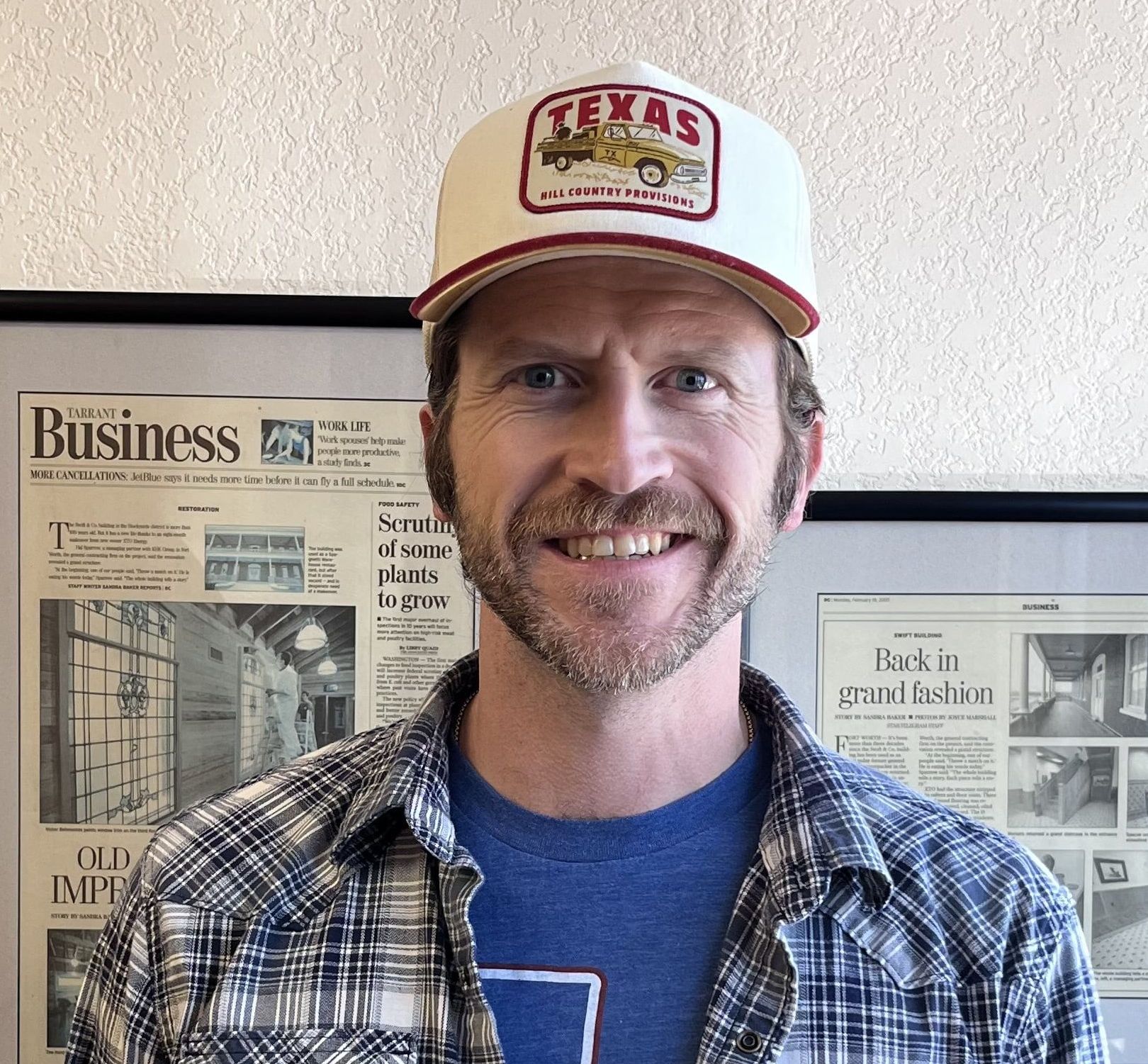 Man wearing a Texas hat, smiling. He has a beard and is in front of newspaper pages.