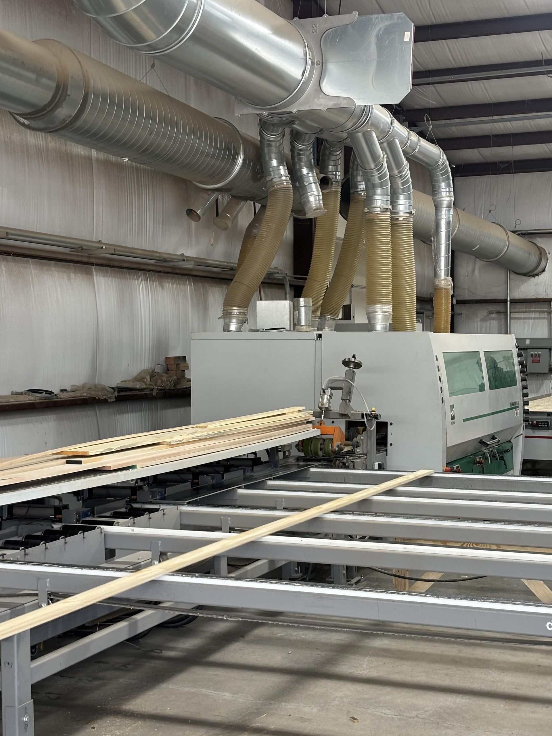 Factory machine processing wood, with ventilation system above.  Long wooden planks are moved along a metal track in a workshop.