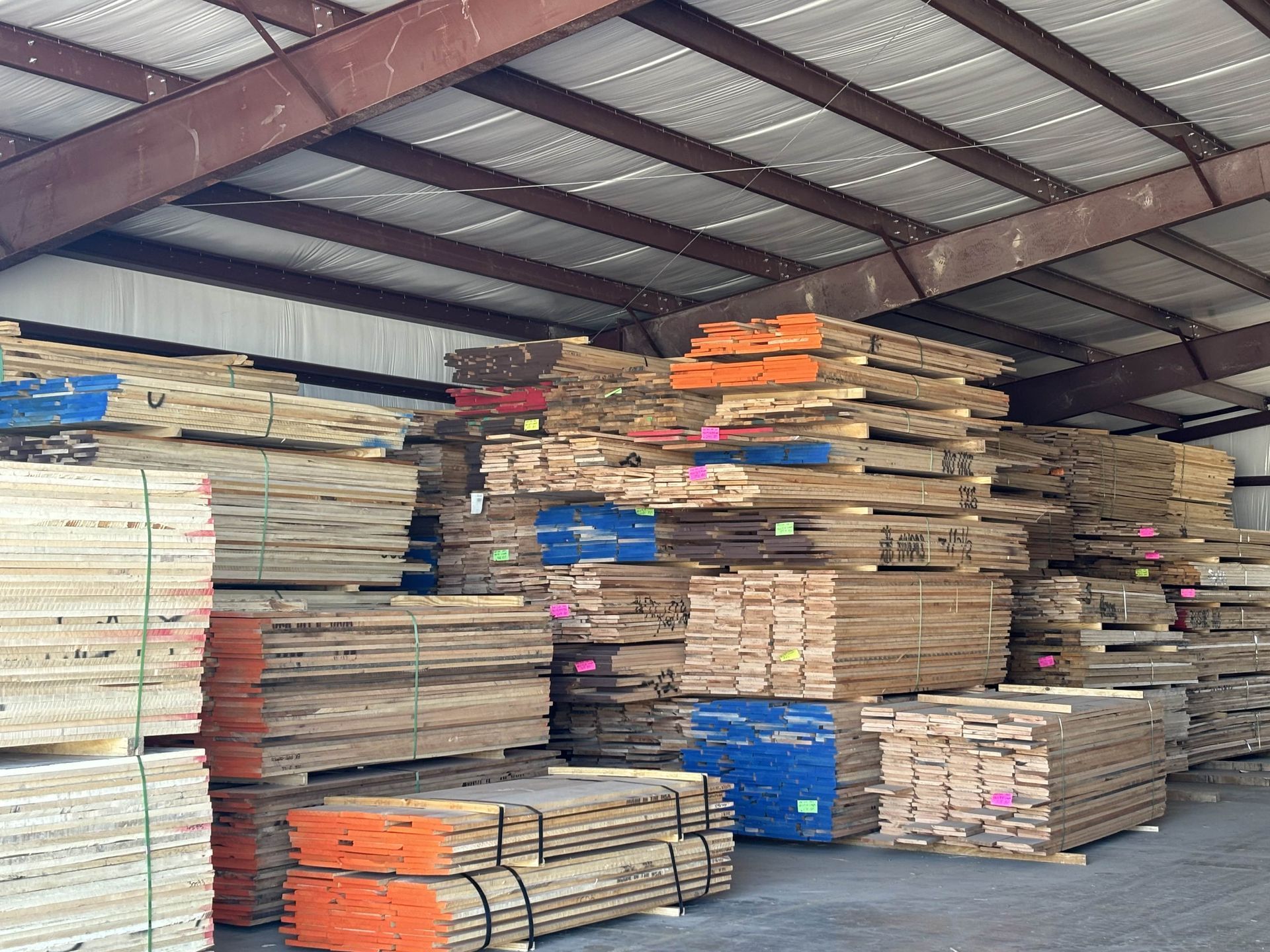 Stacks of lumber in a warehouse. The wood varies in color, with some pieces marked with blue, orange, and pink paint.