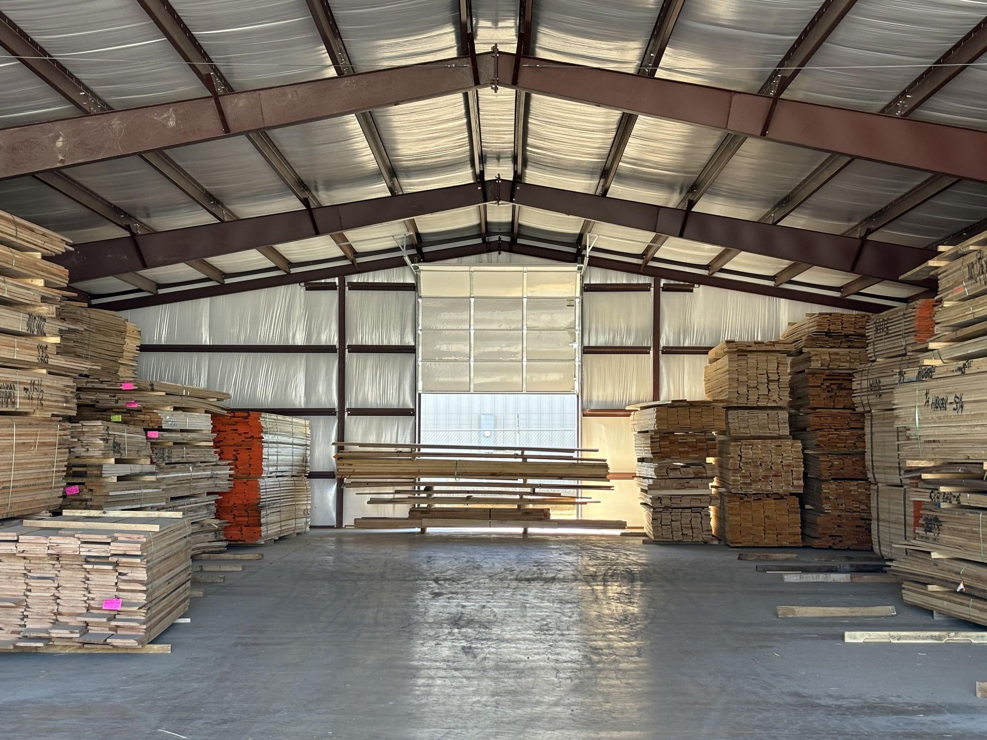 Lumber stacked inside a warehouse. The interior features metal beams, insulation, and a concrete floor with natural light streaming in.