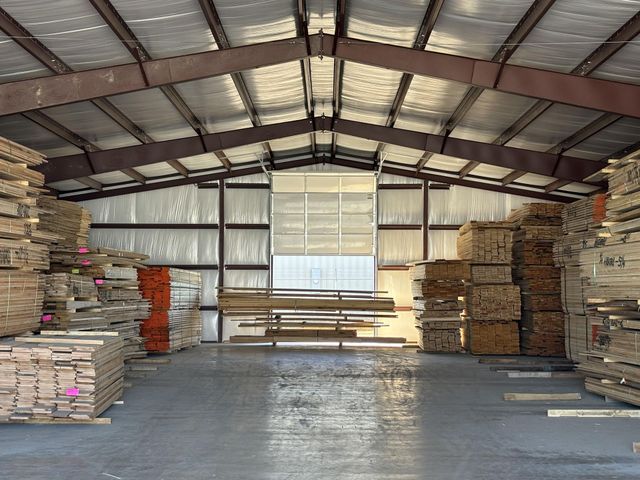 Lumber stacked inside a warehouse. The interior features metal beams, insulation, and a concrete floor with natural light streaming in.