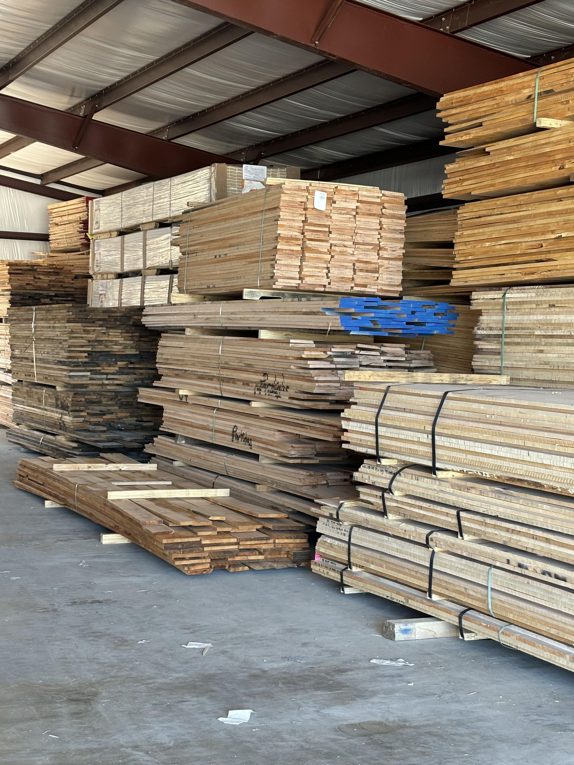 Stacks of lumber stored inside a warehouse. The wood varies in size and color, with many pieces bundled together.