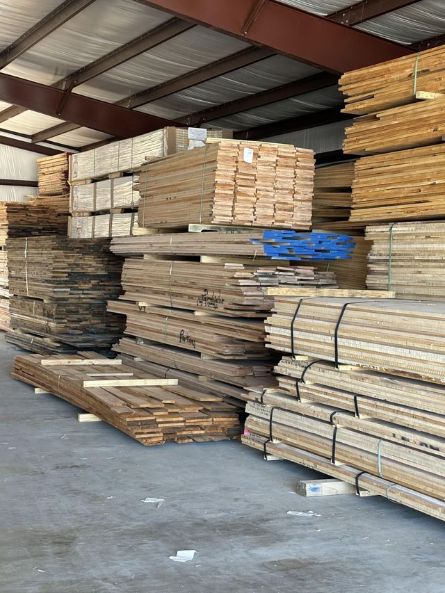 Stacks of lumber stored inside a warehouse. The wood varies in size and color, with many pieces bundled together.