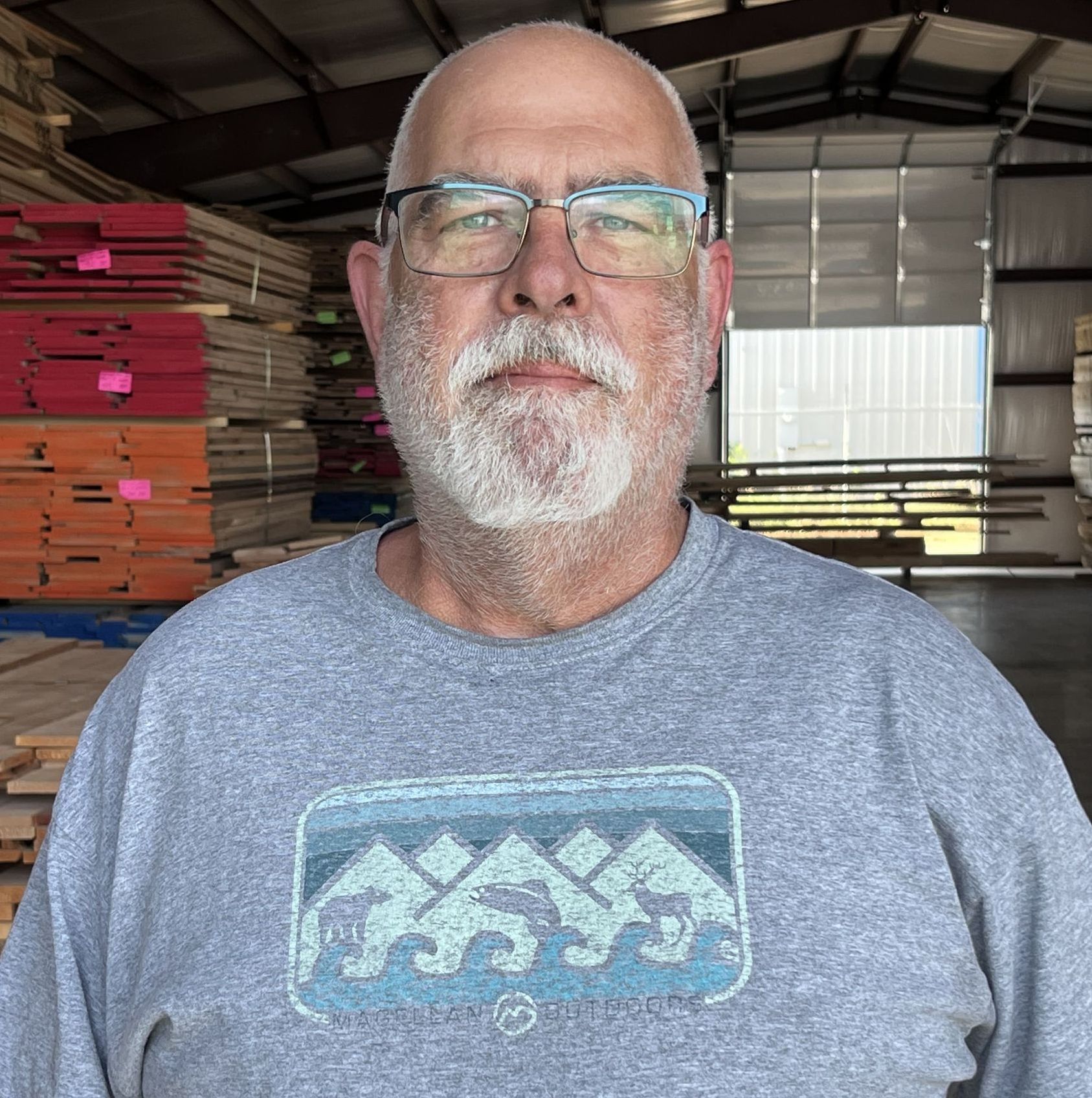 A middle-aged man with a white beard and glasses stands in front of stacks of lumber, wearing a gray t-shirt with a mountain design.