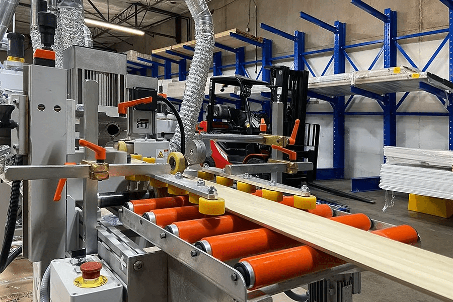 A wood milling machine cutting a long piece of lumber. The scene is set in a factory, with a forklift and shelves in the background.