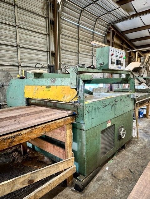 A large, industrial wood planer machine in a workshop. The machine is green and yellow, with a piece of wood being fed into it.