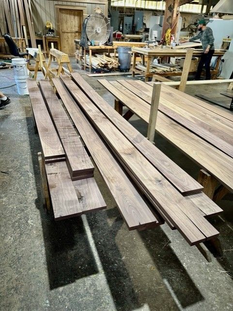 Wooden planks stacked in a woodworking shop, with a worker in the background. Dark wood.