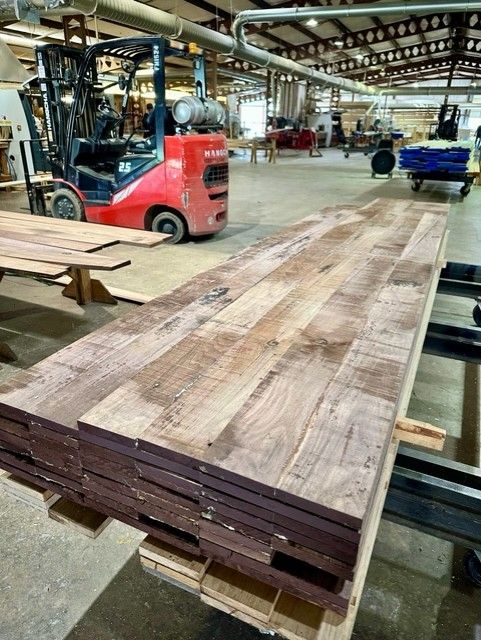 Stack of wooden planks on a pallet in a woodshop, with a forklift in the background. The planks are brown and have visible wood grain.