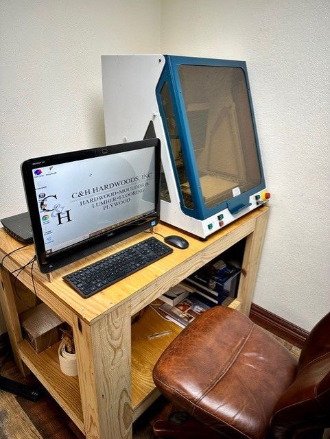 A laser engraving machine and computer setup on a wooden desk in a corner, with a brown leather chair.