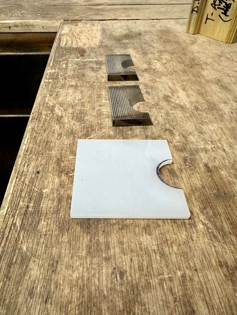 Three pieces of wood and plastic on a wooden surface. The top piece is dark and rectangular. The middle piece is light-colored and ridged. The bottom piece is white and square with a curved cutout.