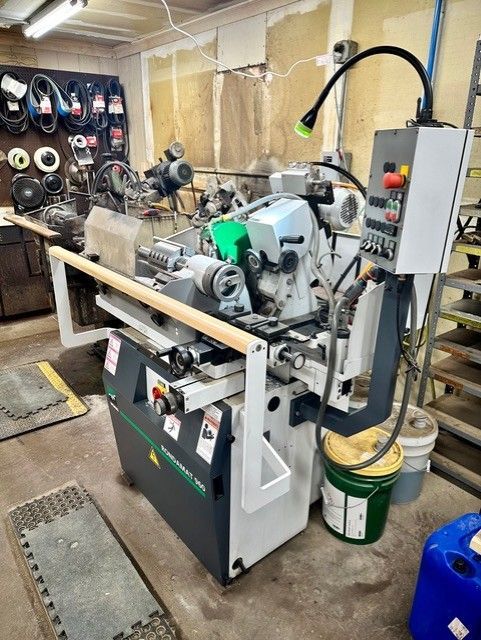 A metalworking machine in a workshop, with a control panel and attached light. The machine is grey, green, and white, with some tools in the background.