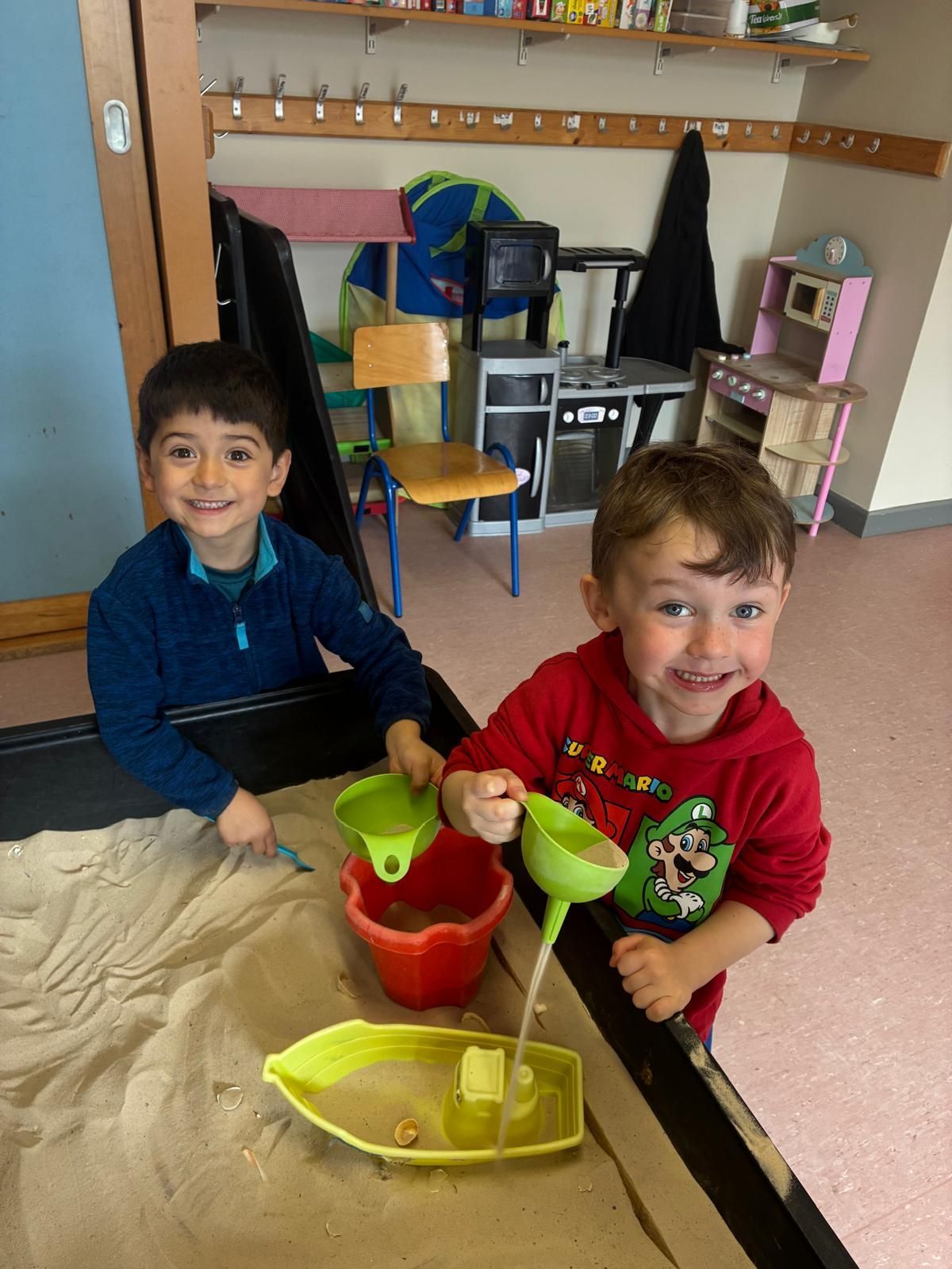 Two children playing with toy dishes in a classroom sand table, smiling at the camera.