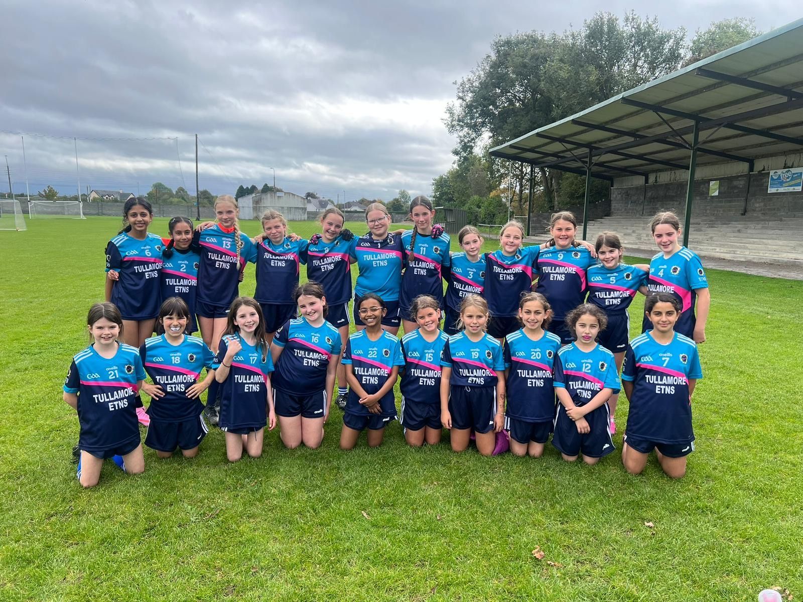 Youth sports team posing on a grassy field beside a covered stand under cloudy skies.