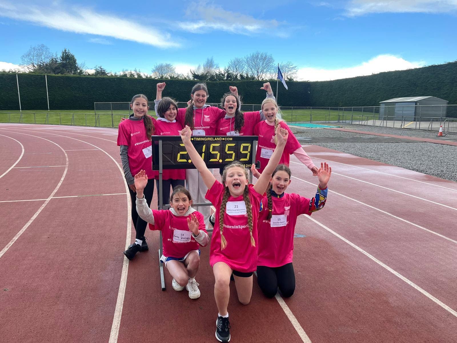 Six young athletes in pink jerseys pose on a track with a finish banner, smiling and raising their arms.