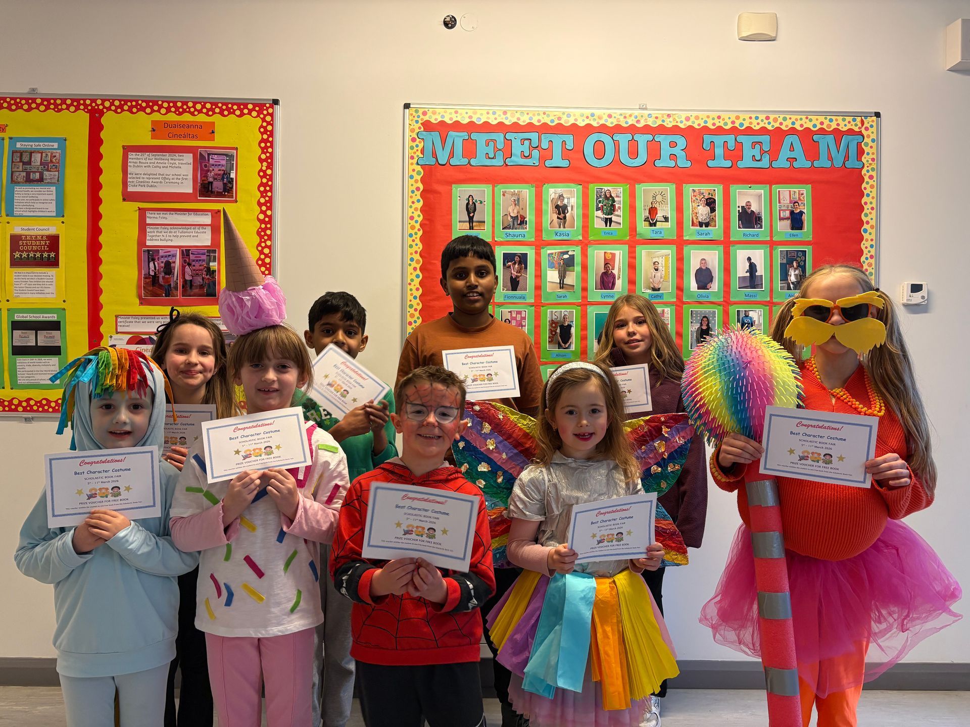 Children posing with certificates in a classroom display, with a “Meet Our Team” bulletin board behind them.