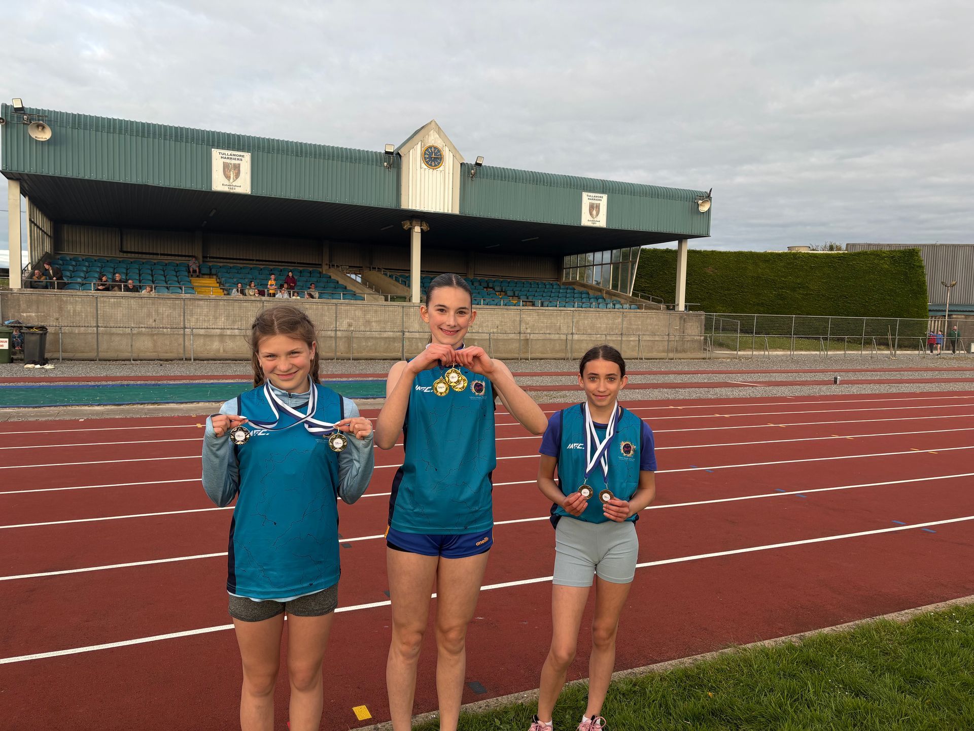 Three medal-winning athletes stand on a track podium holding medals, with a stadium in the background.