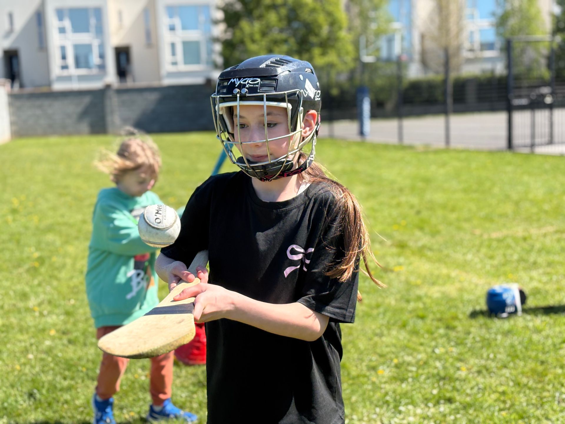 Girl in a black helmet catching a ball on a grassy field, with a child in green behind her.