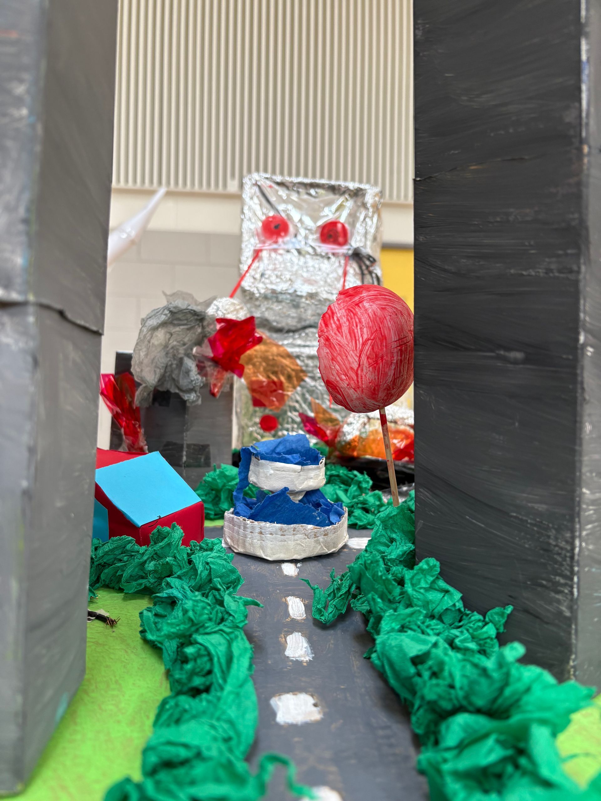 Miniature model city with a gray road, green trees, and a foil robot figure with red eyes.