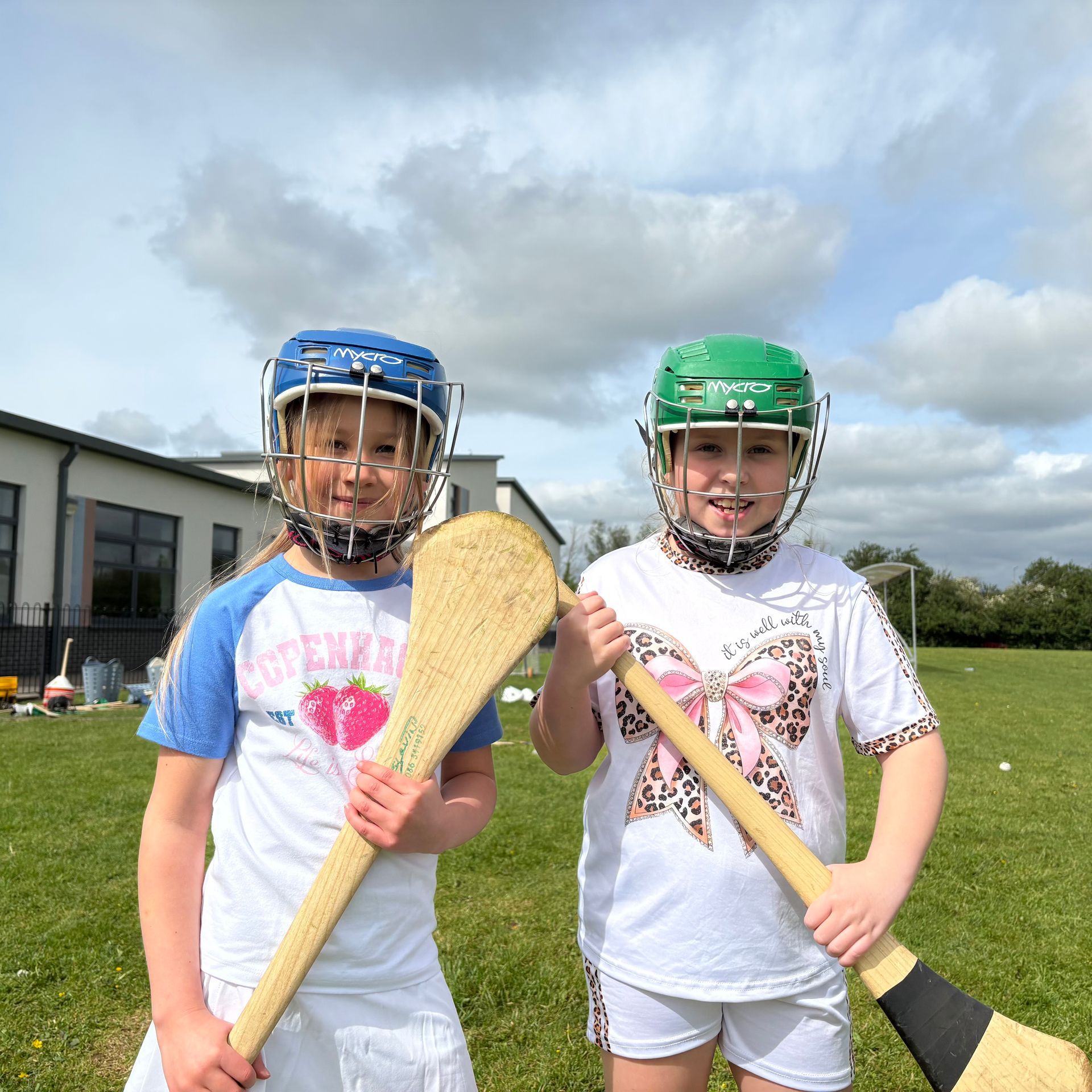 Two children in helmets hold hurley sticks on a grassy field under a cloudy sky.