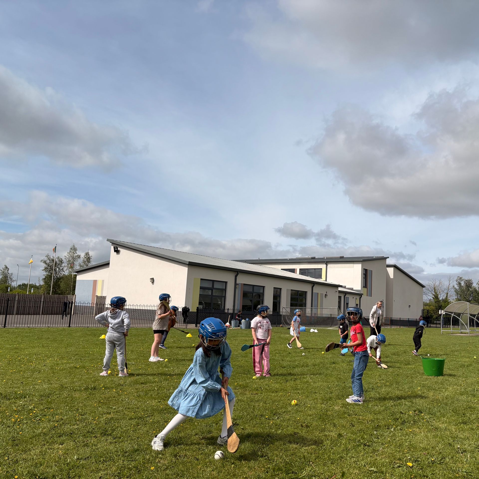 Children playing outdoors on a grassy field near a school building, with a cloudy sky overhead.
