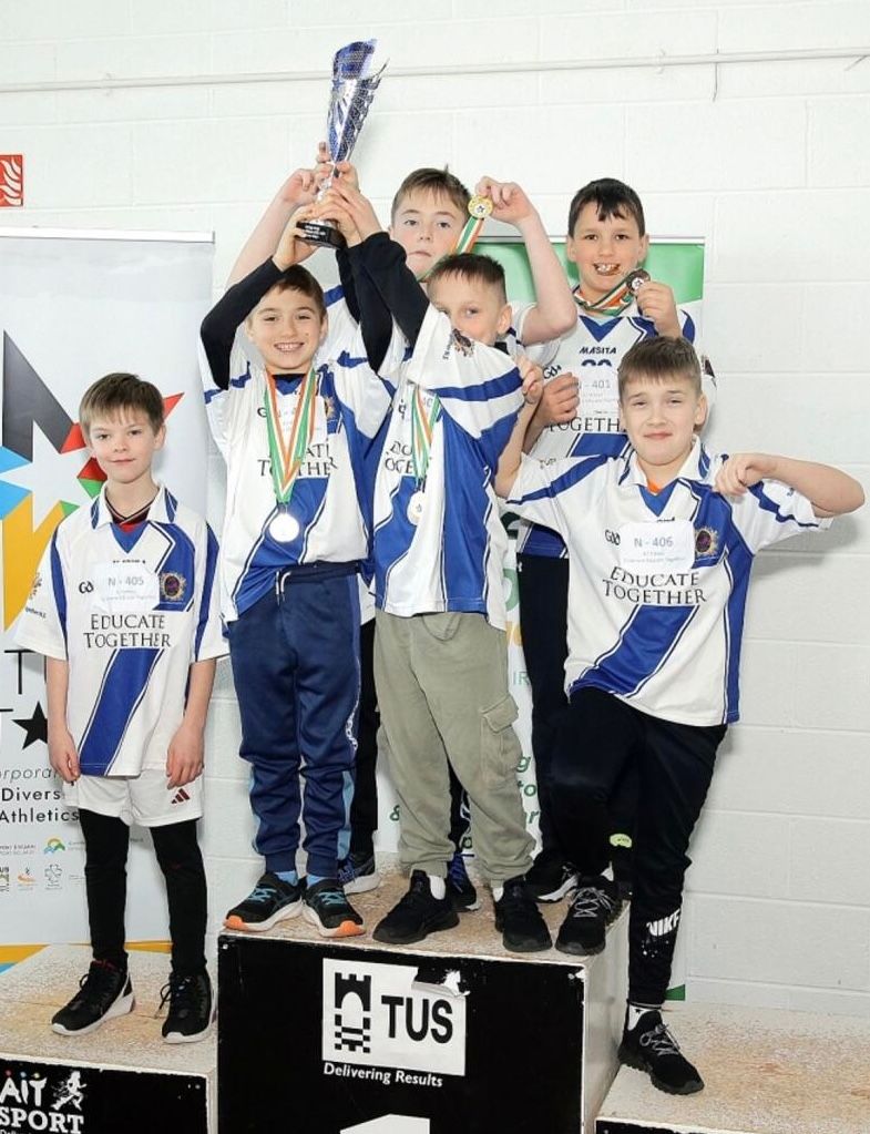 Children in white and blue sports uniforms celebrating on a podium, one holding a trophy.