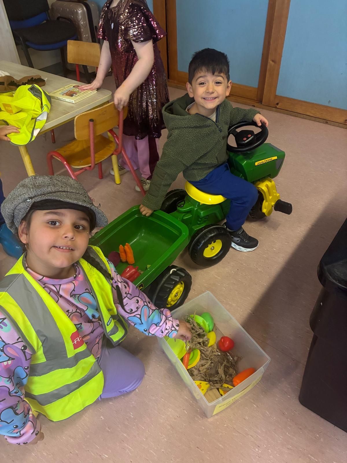 Children playing indoors with a green toy ride-on tractor and a clear bin of colorful toys