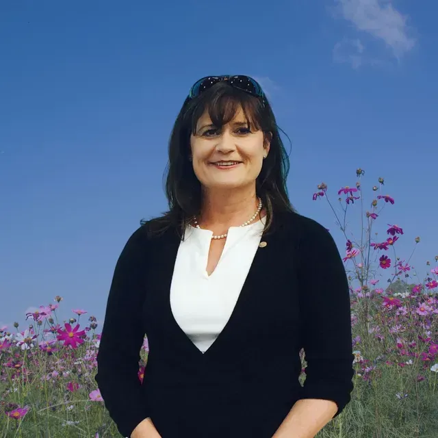 A woman in a black sweater and white shirt is standing in front of a field of flowers.