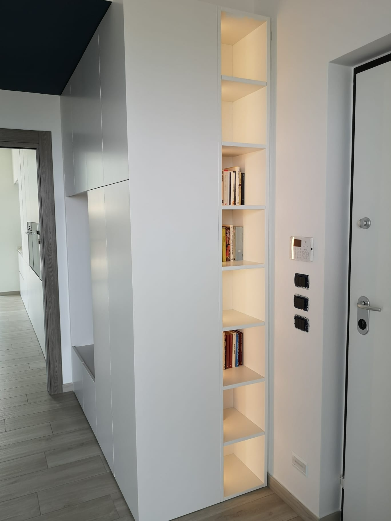 Libreria bianca stretta con illuminazione interna accanto a una porta bianca in un corridoio.