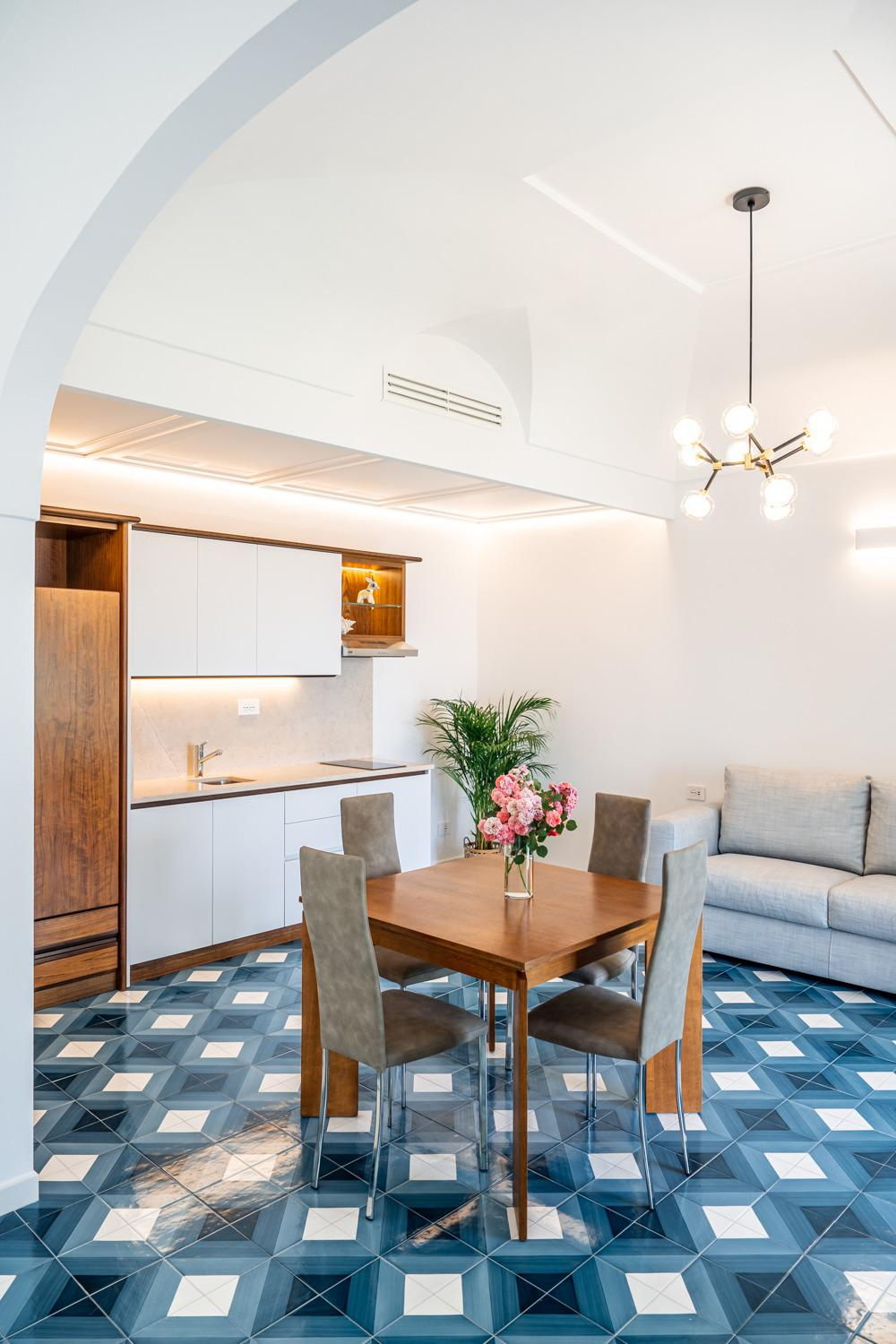 Appartamento open space con pavimento in piastrelle blu, cucina, tavolo da pranzo con sedie e divano.