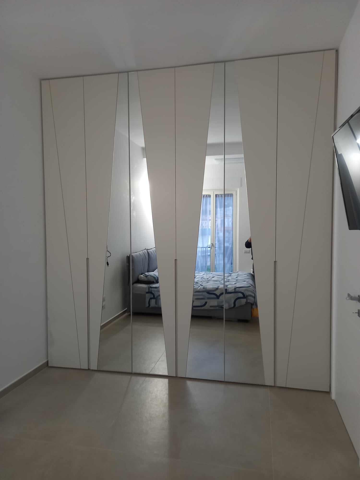 Le ante bianche dell'armadio con pannelli a specchio riflettono la camera da letto con letto e divano.