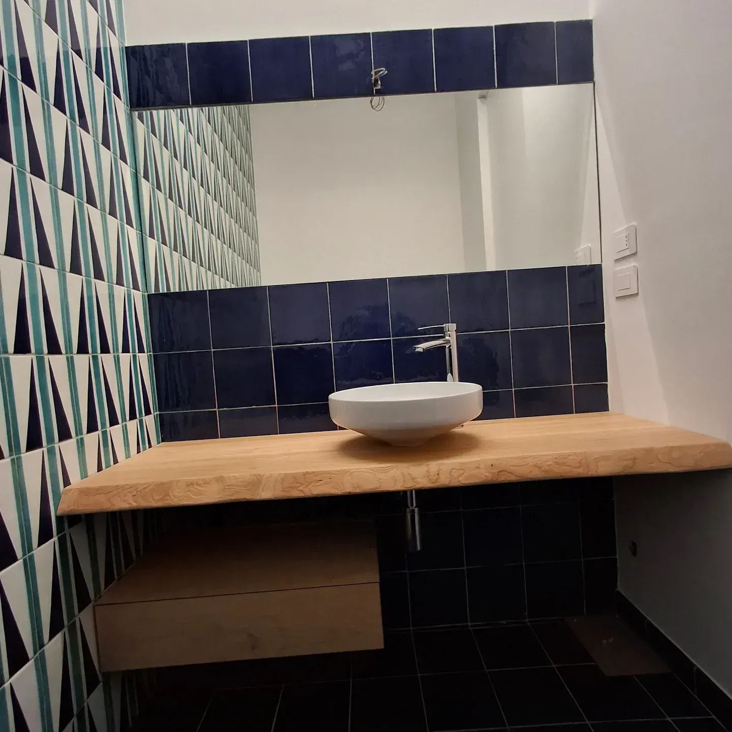 Bagno con piastrelle a motivi blu e verde acqua, un bancone sospeso in legno, un lavandino bianco e un grande specchio.