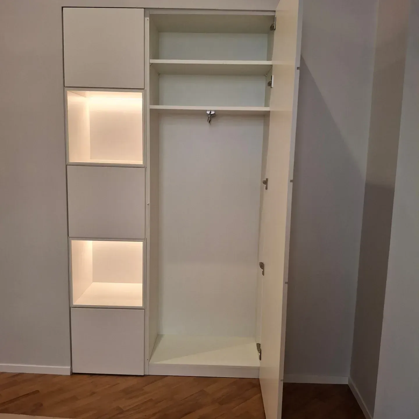 Armadio bianco a muro con ripiani e asta appendiabiti; sul lato sinistro sono presenti ripiani illuminati e porta aperta.