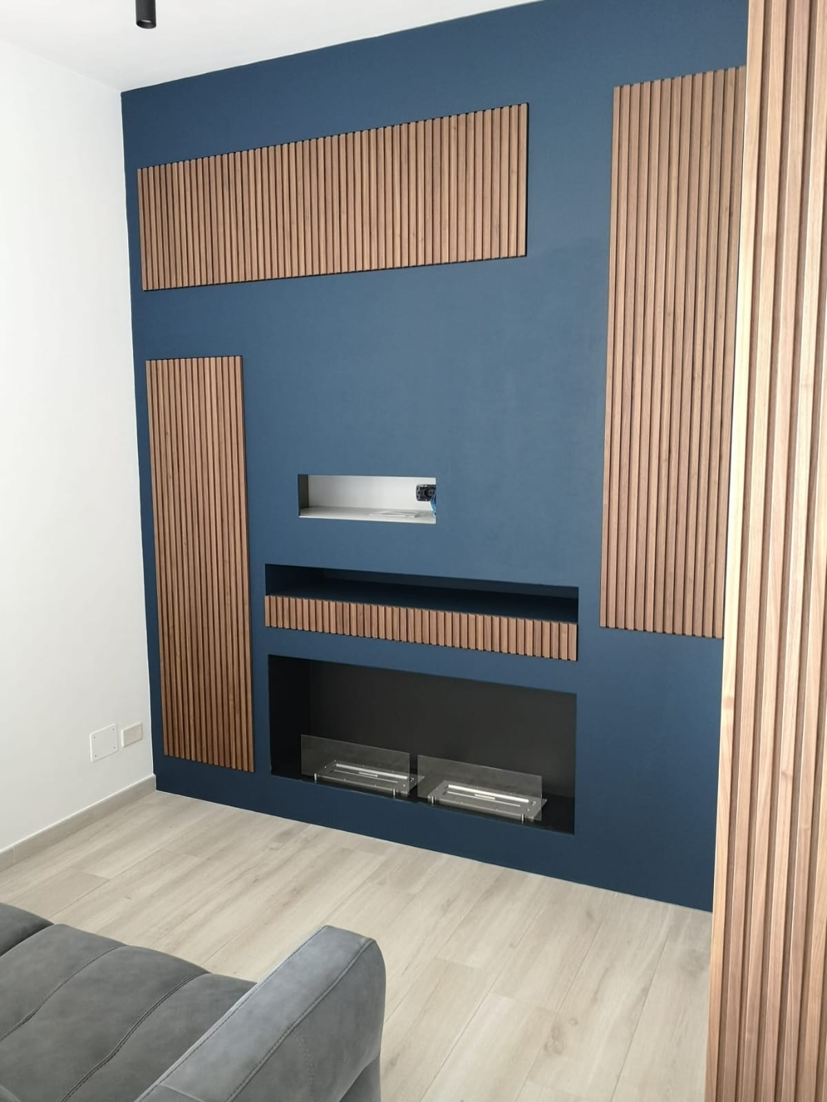 Parete blu con pannelli in legno e camino. Divano grigio in primo piano, pavimento in legno chiaro.