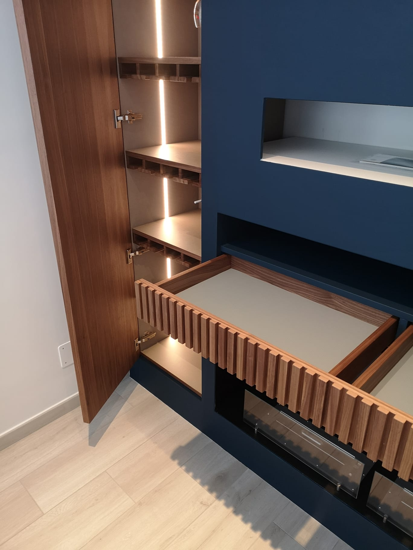 Mobiletto in legno con anta aperta che rivela ripiani e cassetto integrato con frontale in legno testurizzato.