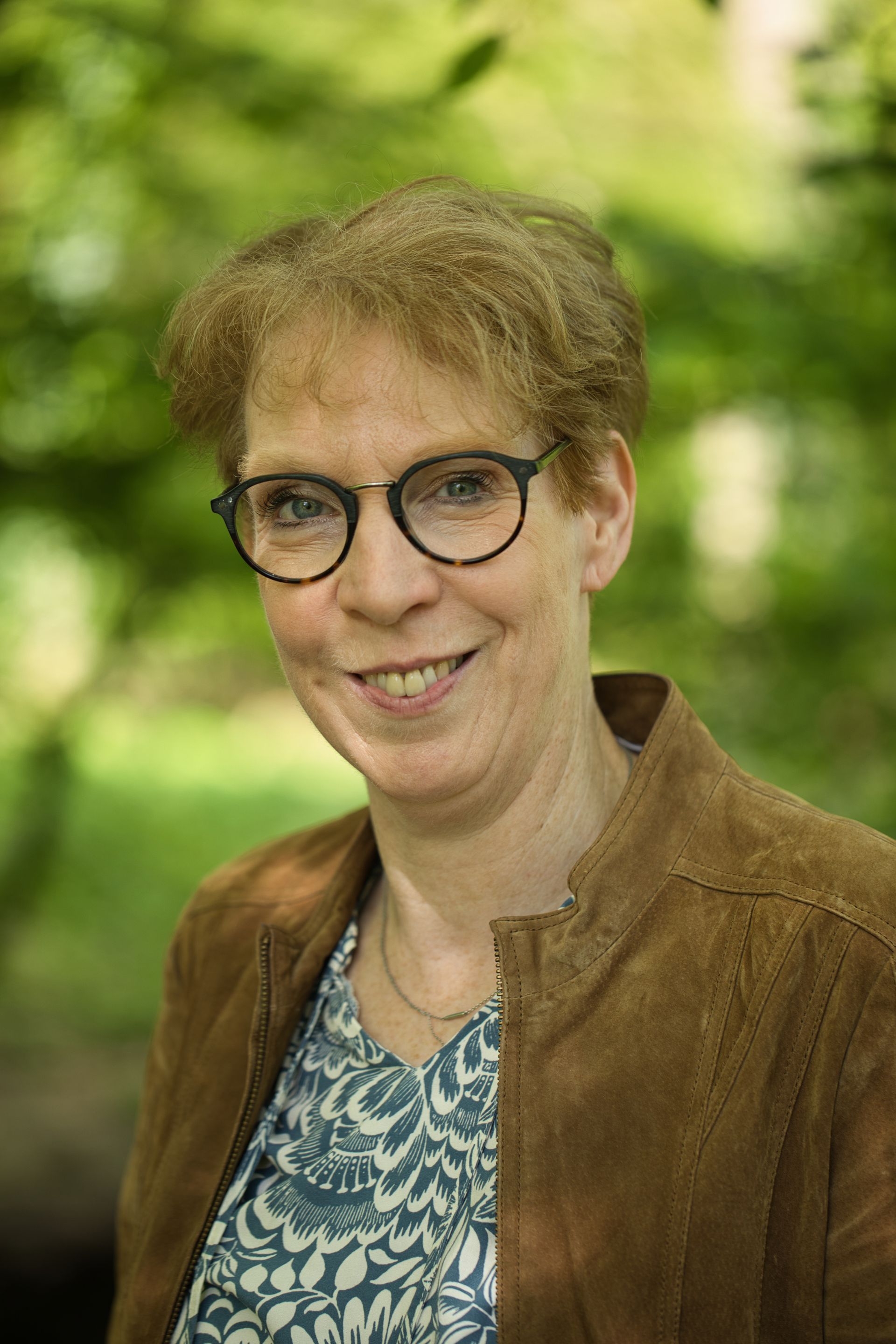 Vrouw met bril en bruine jas lacht buiten.