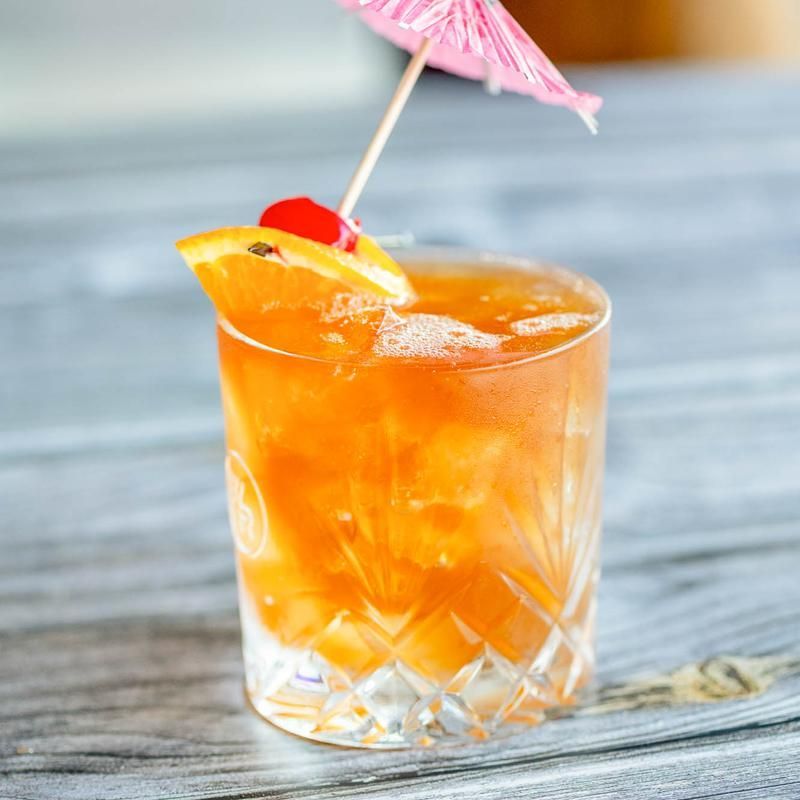 Orange cocktail in a short glass with cherry, lemon wedge, and pink umbrella on a wooden table