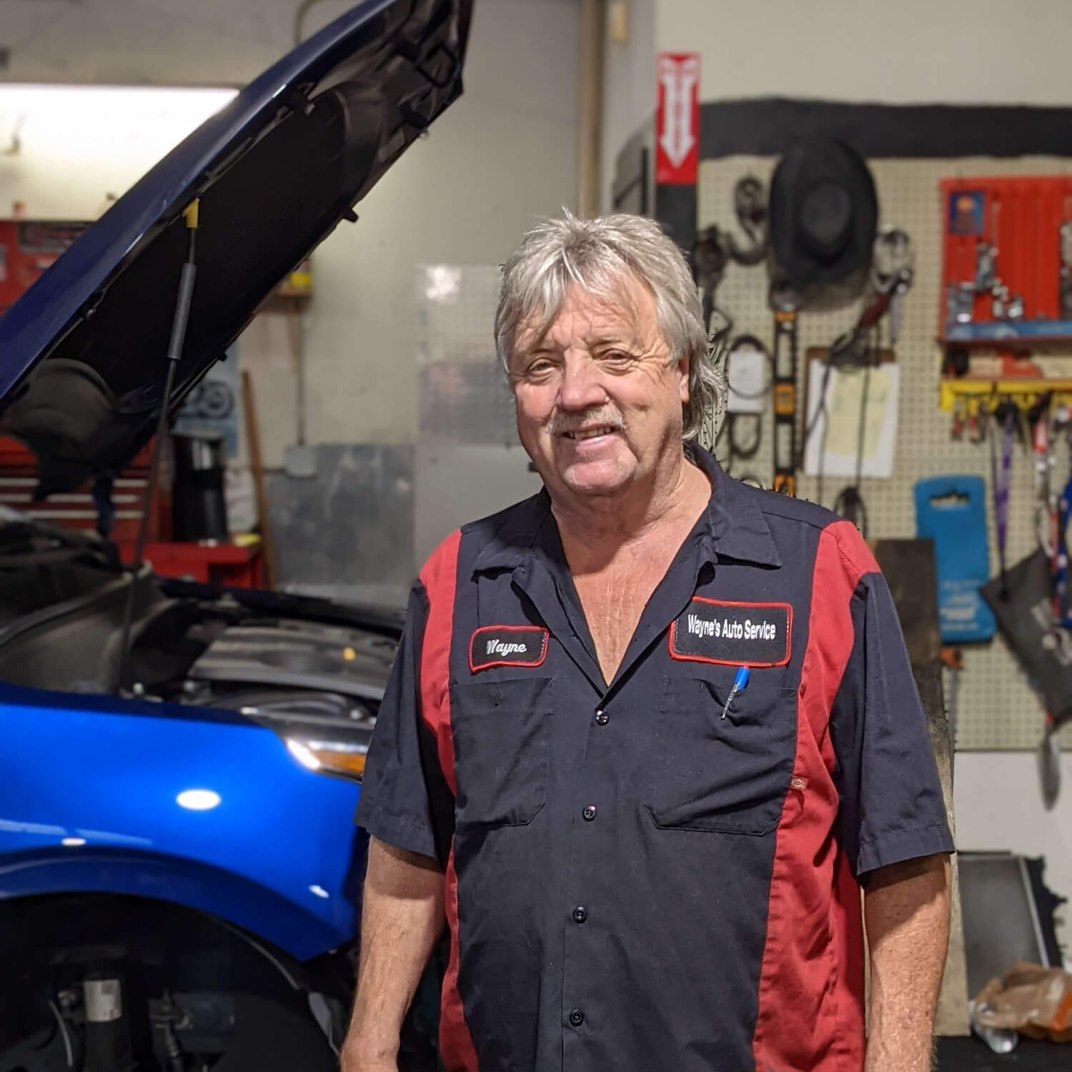 Mechanic in a shop, stands near a blue car with hood up. Wearing a uniform, smiling. Tools and equipment visible.
