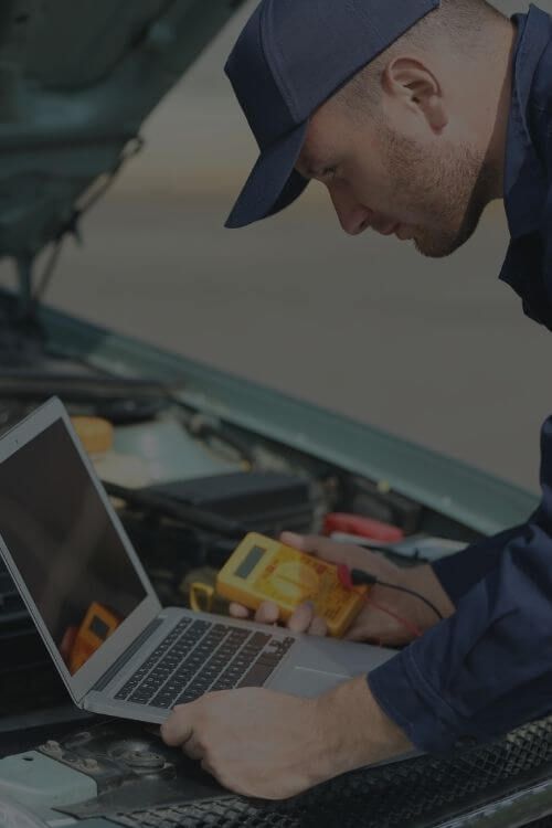 Mechanic using a laptop and multimeter on a car engine.