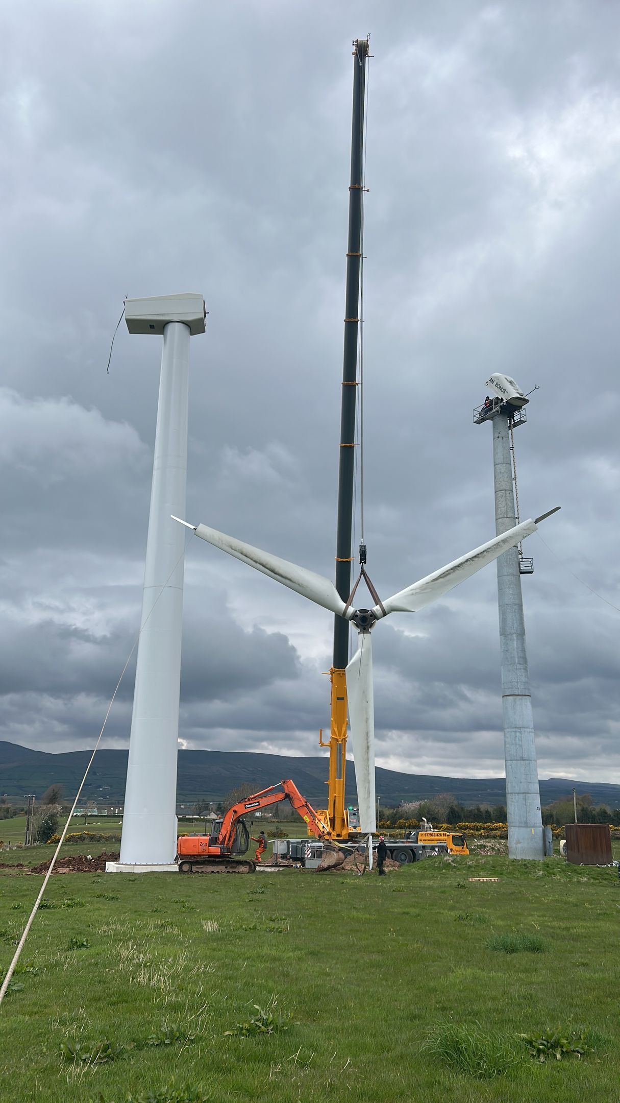 Wind turbine assembly site with a crane, white turbine towers, and workers on a grassy field under cloudy skies