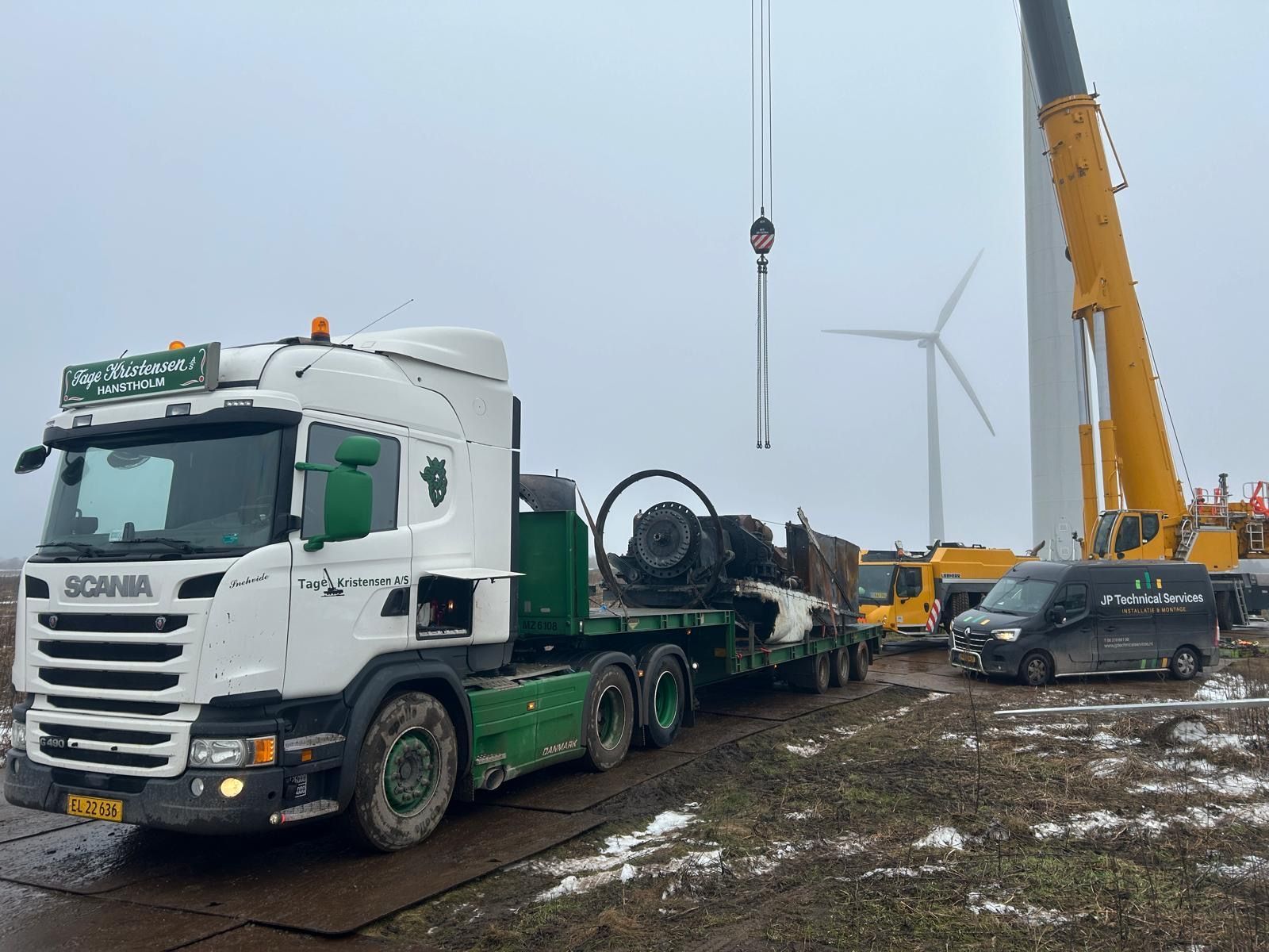 Heavy truck and crane at a muddy wind farm, with a utility vehicle and turbines in the background