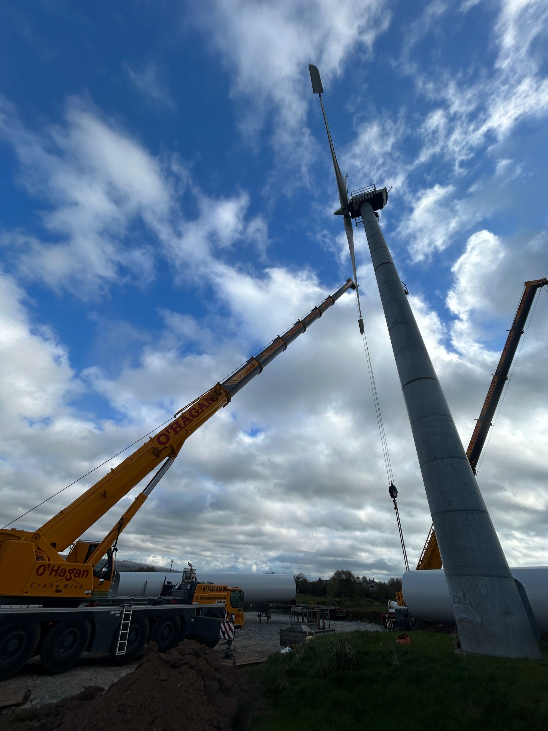 Two cranes lifting a tall gray tower against a cloudy blue sky