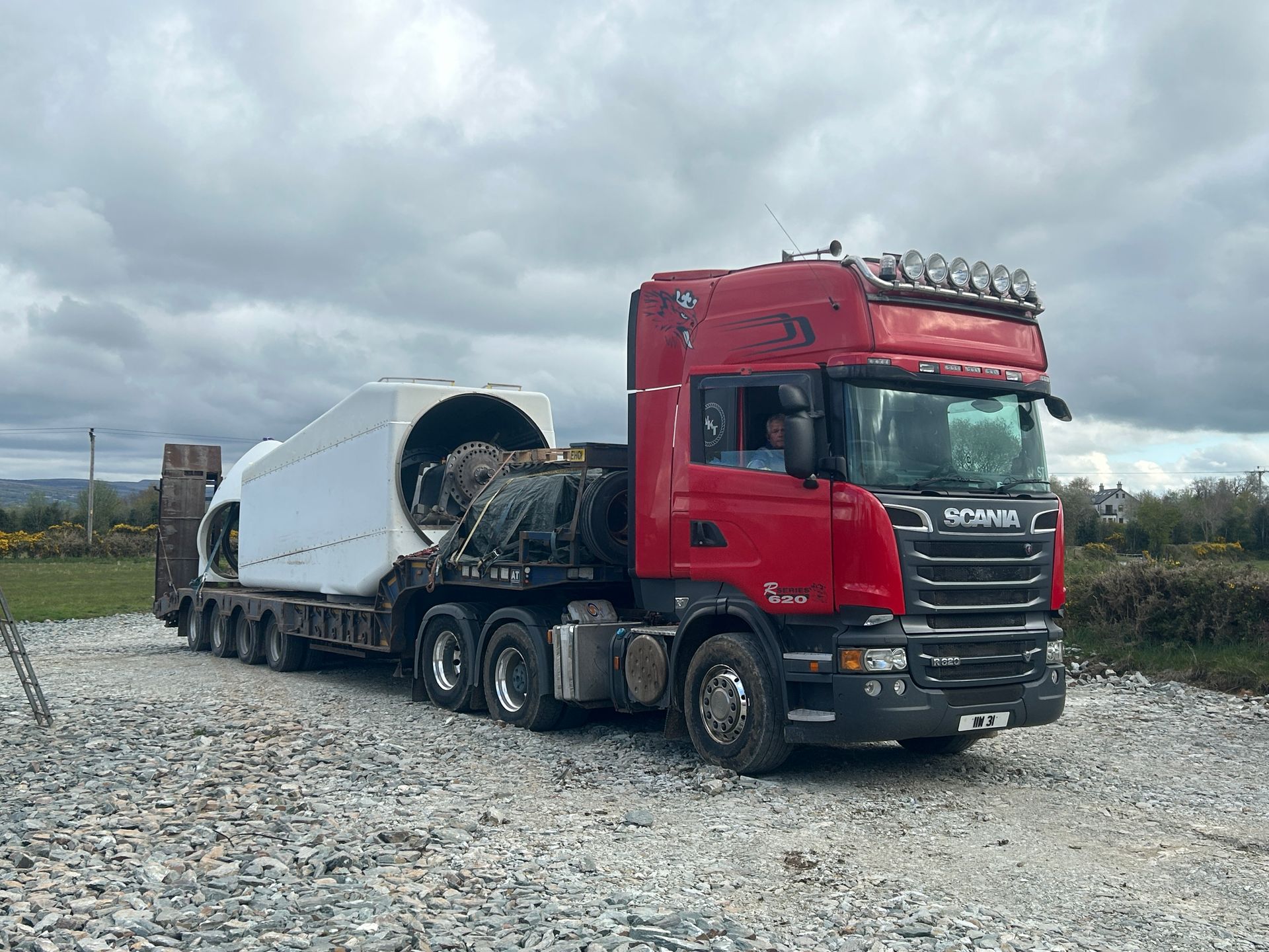 Red semi-truck hauling a large white cylindrical load on a gravel lot under cloudy skies