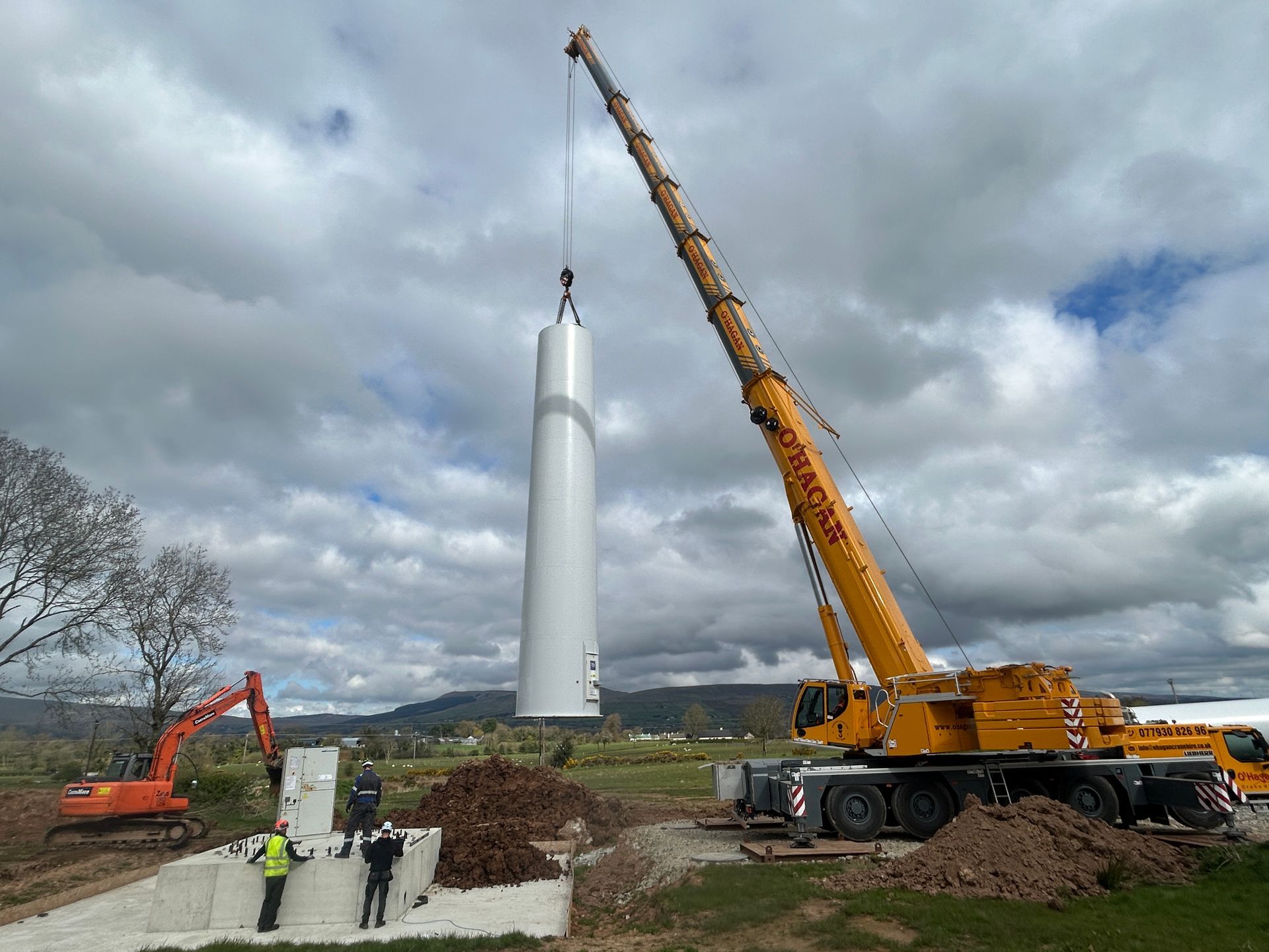 Workers and cranes erecting a tall white wind turbine tower under cloudy skies