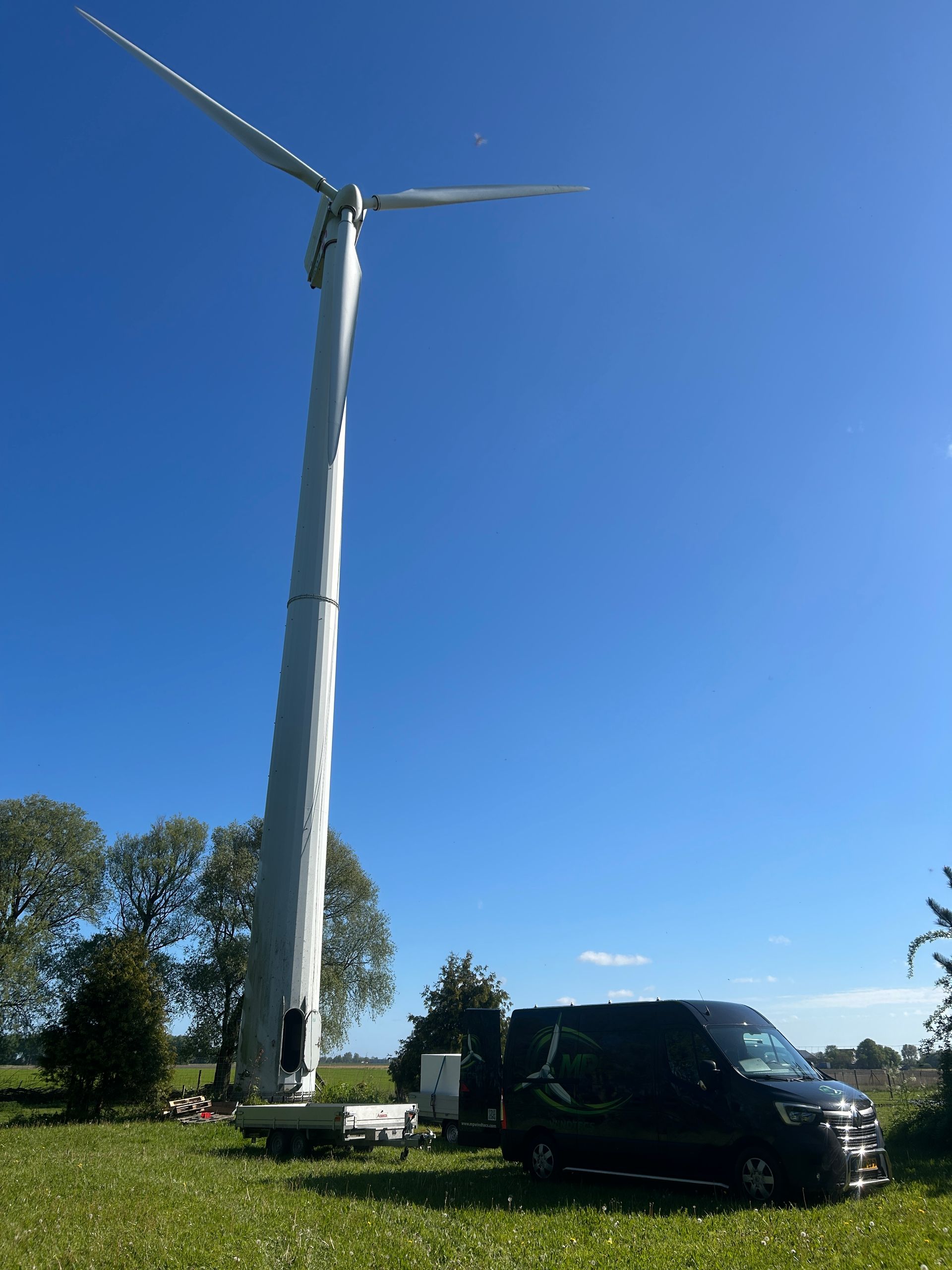 Wind turbine tower beside a black van in a sunny open field under a clear blue sky
