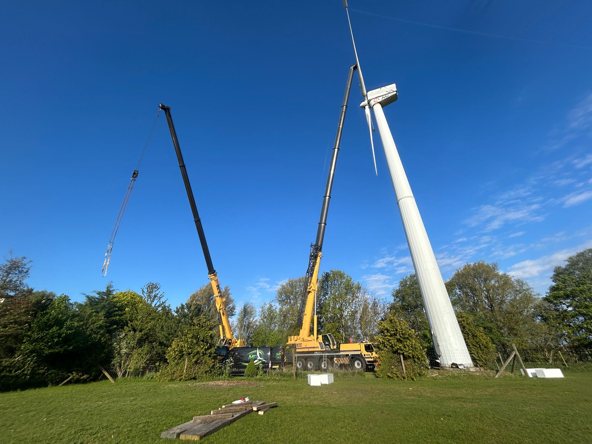 Two yellow cranes and a tall white mast against a blue sky on a grassy field